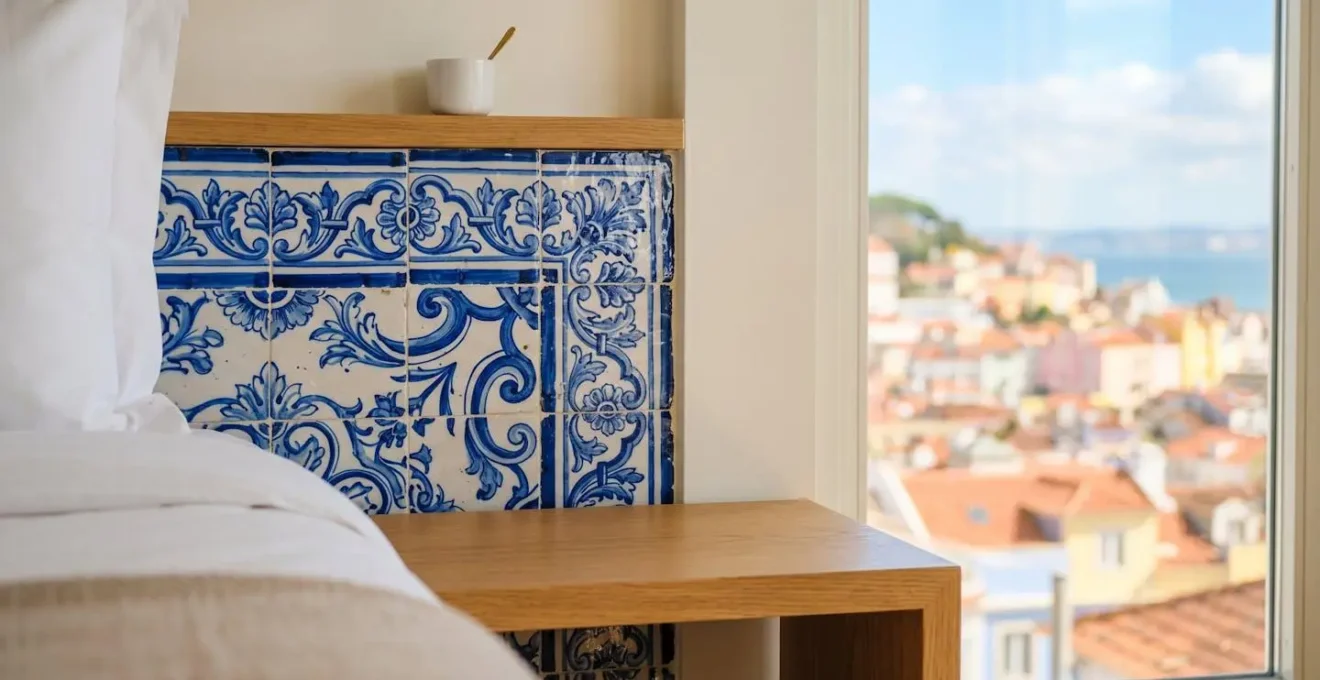 Intérieur lumineux d'une chambre d'hôtel boutique lisboète avec azulejos bleus traditionnels au mur, mobilier design contemporain minimaliste et grande fenêtre donnant sur les toits colorés de la ville